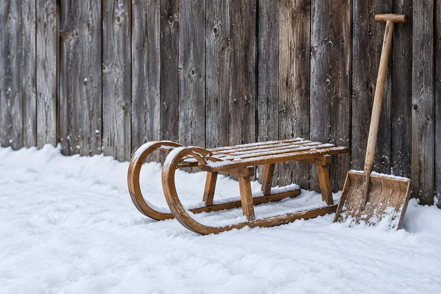 Holzschlitten und Schneeschaufel im Schnee vor einer Holzwand
